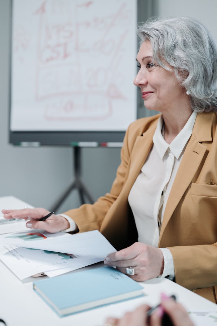 Elderly woman in a blazer at a meeting, holding documents, with a presentation board in the background.